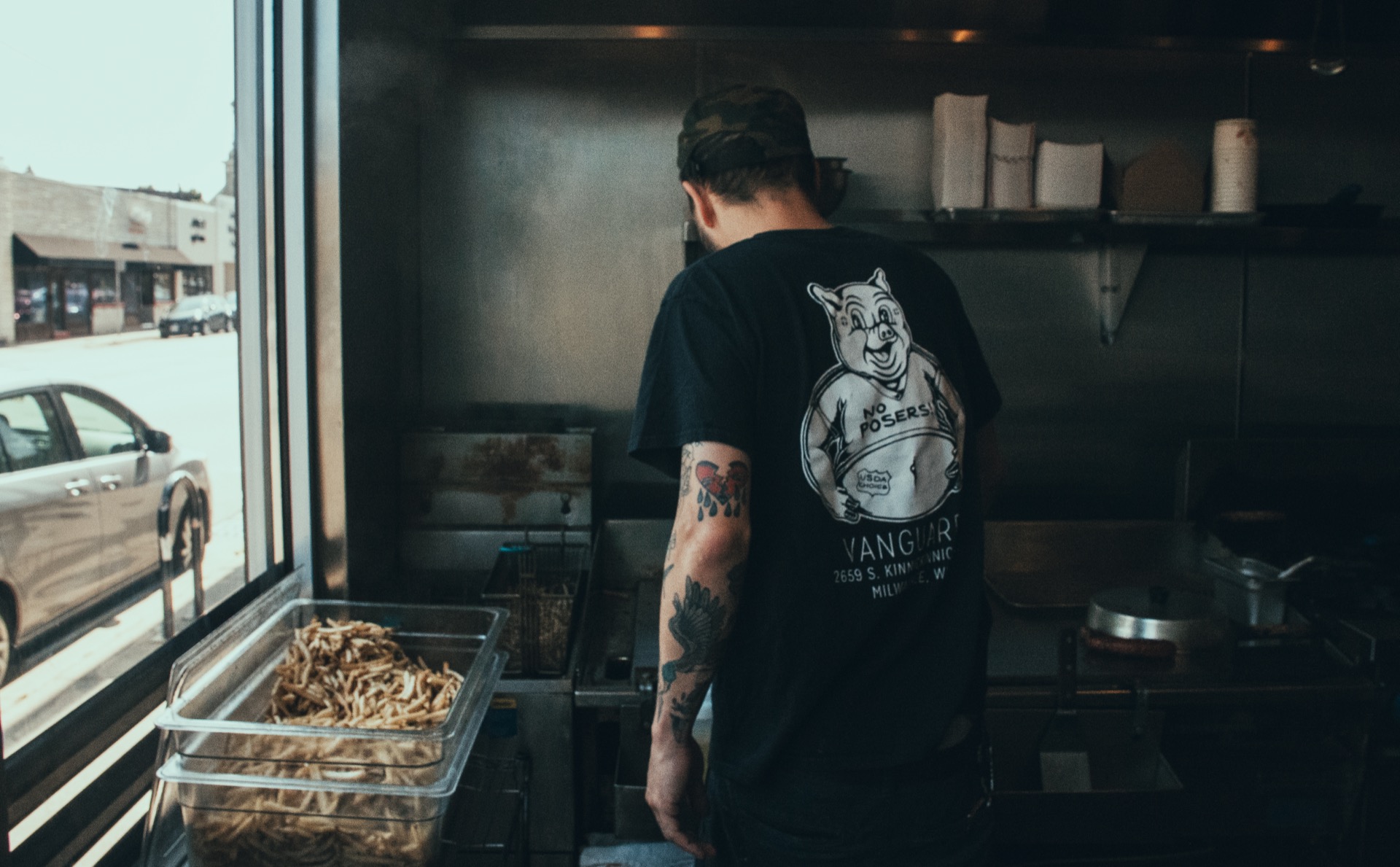 Tattooed cook in a Vanguard t-shirt working the fryer by the window